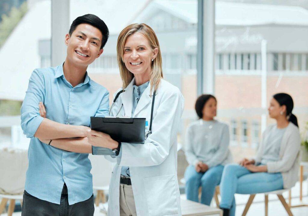 A male healthcare worker and a female doctor smiling at the camera with patients seated in the background.